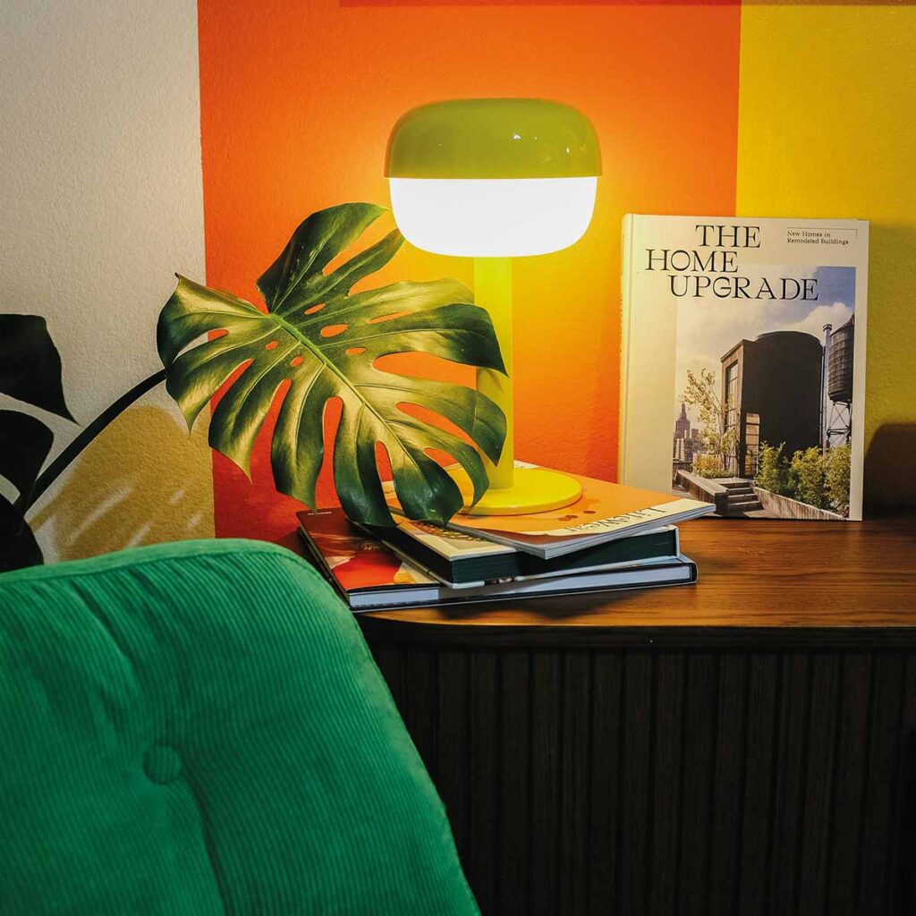 Interior-Styling mit grüner Tischleuchte, Coffee Table Books, Monstera-Blatt und kräftigen Farbakzenten in Orange und Gelb