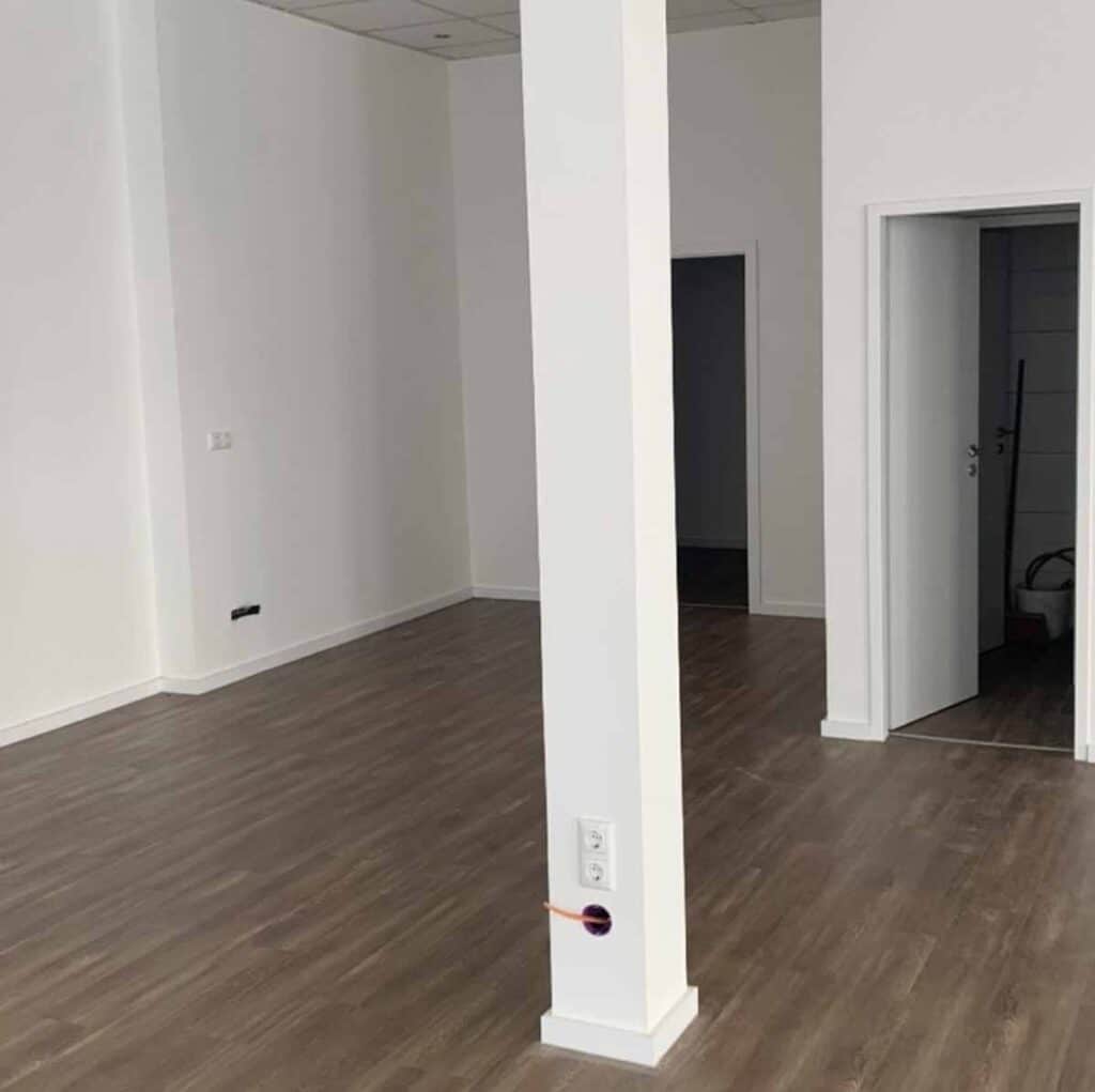 Leere Ladenfläche im Rohzustand mit weißen Wänden, dunklem Vinylboden, einer tragenden Stütze mit Steckdose und Türen zu den Nebenräumen.
