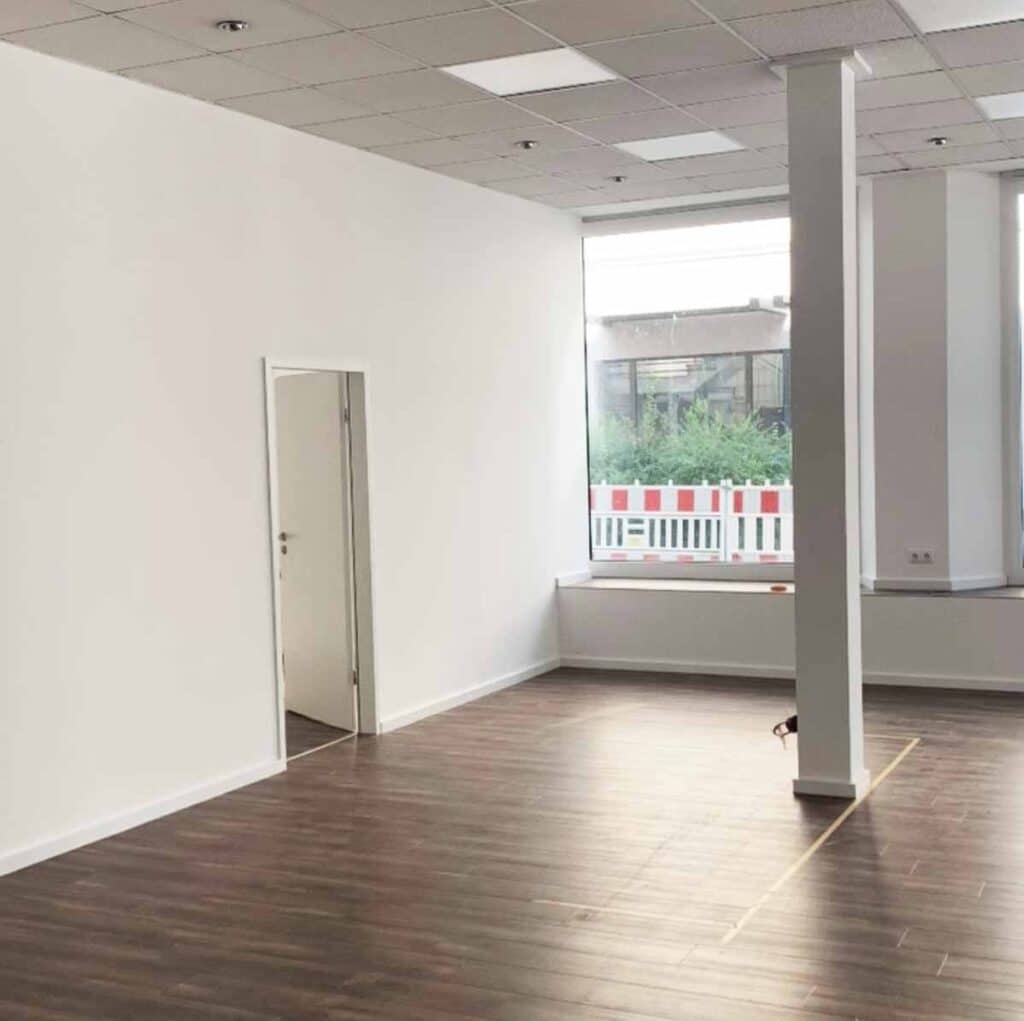 Leerstehendes Ladengeschäft im Rohzustand mit weißen Wänden, Rasterdecke, dunklem Vinylboden und einer tragenden Stütze vor dem Schaufenster.